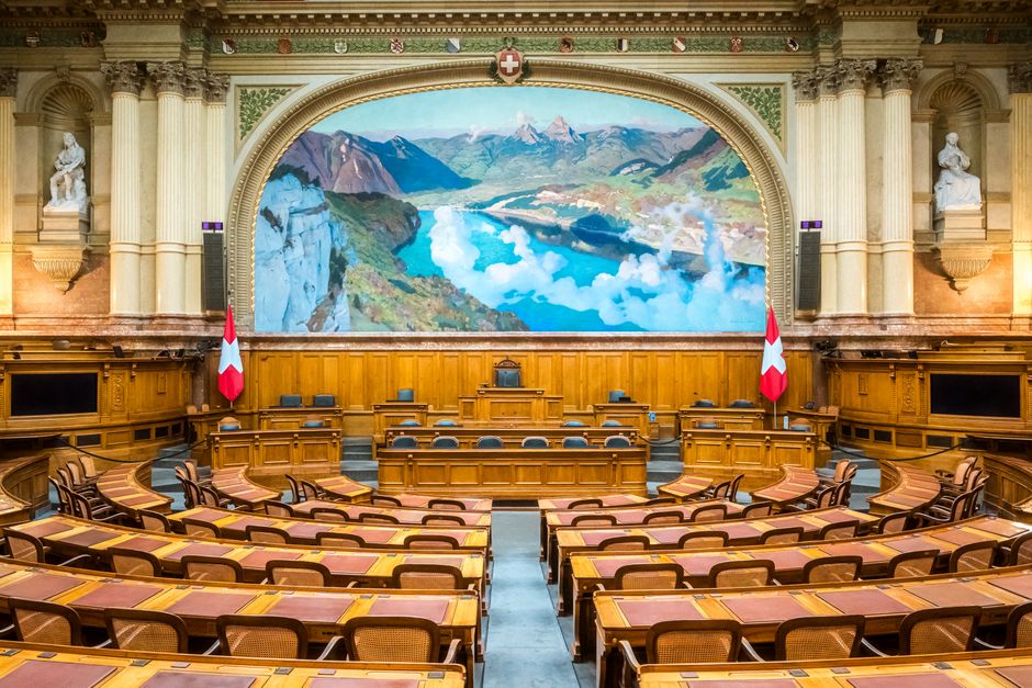 A black-and-white photograph showing rows of wooden desks equipped with microphones and glasses of water in a large, historic council chamber or courtroom. The desks are arranged in parallel lines, wi