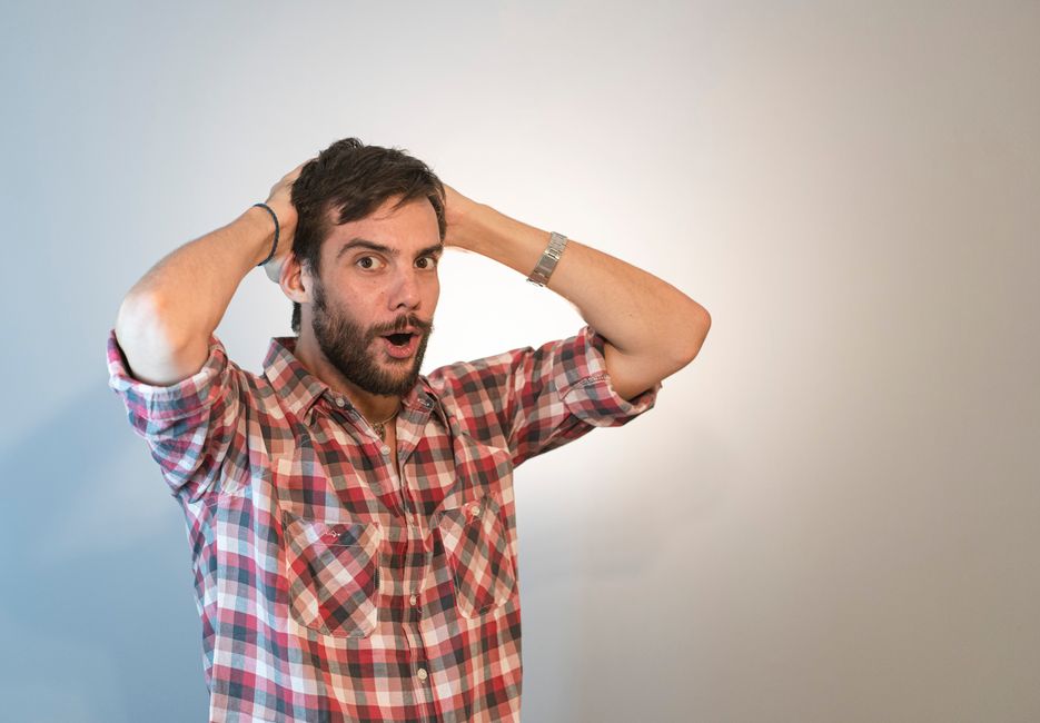 A man with dark hair and a beard, wearing a red, white, and blue checkered shirt, stands indoors against a plain, light-colored background with natural lighting. He has a surprised or puzzled expressi