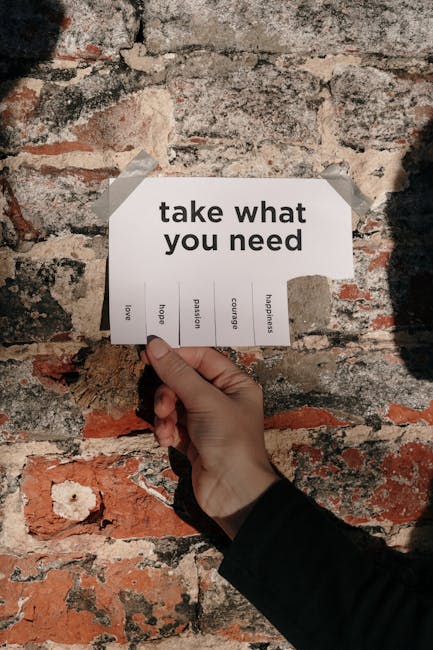 A person wearing a dark long-sleeve garment is holding a white paper card with black printed text that reads 'take what you need' and several smaller printed words such as 'love,' 'hope,' 'passion,' '