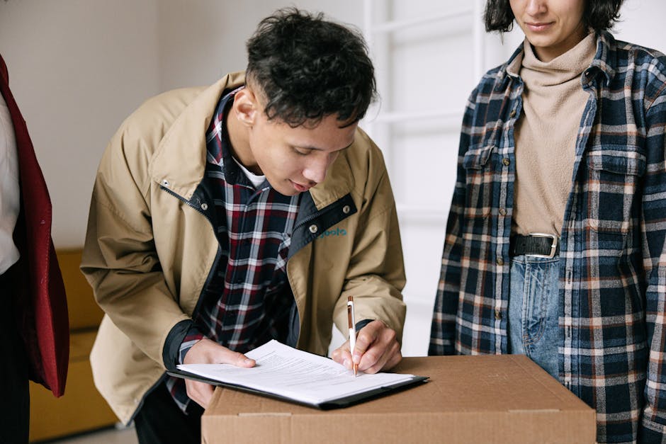 A young man with curly dark hair, wearing a beige jacket over a red, blue, and white checkered shirt, is carefully signing a document on a clipboard. He is leaning over a large cardboard box placed on a wooden surface inside a property. Next to him, a woman with shoulder-length dark hair, dressed in a blue and brown plaid shirt over a beige turtleneck, stands attentively. The background shows a white interior wall with a shelf and natural lighting illuminating the scene, indicative of a house removal or packing process. This image captures the detailed action of a home relocation, focusing on the organization and documentation involved in furniture transport and packing, representative of services offered by removalspaddington.com.