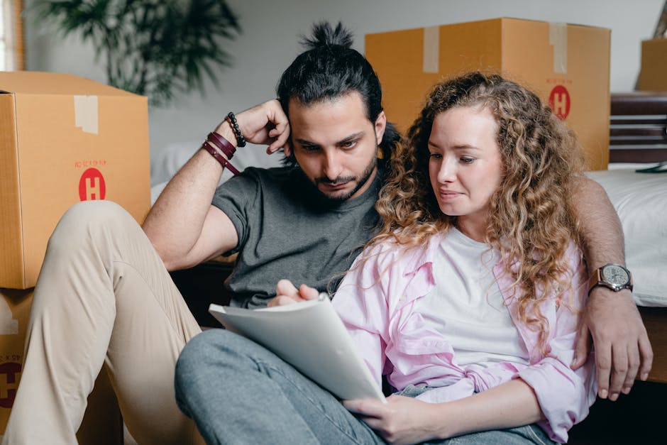 A young man with dark hair tied back and a beard, wearing a grey t-shirt and beige pants, sits on the floor inside a property with his back against a bed. He is holding his head with one hand and looking down at a tablet device being held by a young woman with long, curly blonde hair, who is dressed in a light pink shirt and grey jeans. Surrounding them are several packed cardboard moving boxes with red markings, some sealed with packing tape, indicating that they are in the process of packing for a home relocation. The scene shows the interior of a room with a white wall, and a wooden dresser partially visible on the right. The environment is well-lit, suggesting daytime, and the activity reflects the packing and moving preparations associated with house removals, as handled by [COMPANY_NAME], for a target page about the Norfolk Square move checklist for Paddington residents.